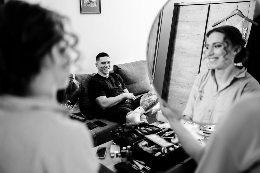 Black and white photo of a man relaxing on a sofa while two women chat nearby with makeup supplies on a table.