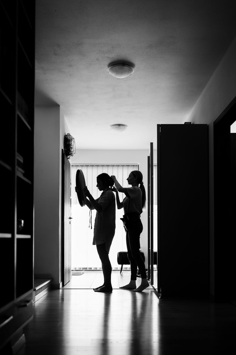 Black and white silhouette of two people standing in a hallway doorway, one holding up a shoe.