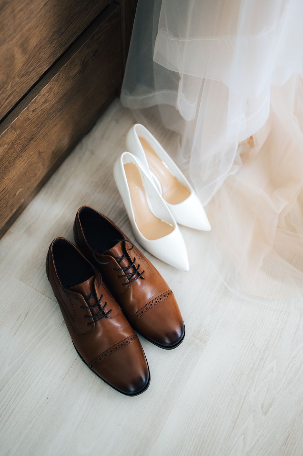 Brown leather oxford shoes and white bridal heels on a light wood floor, perfect for a wedding day.