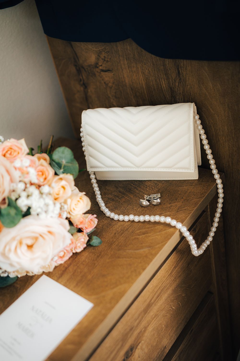 White quilted clutch with pearl strap beside blush roses and wedding invitation on wooden dresser.