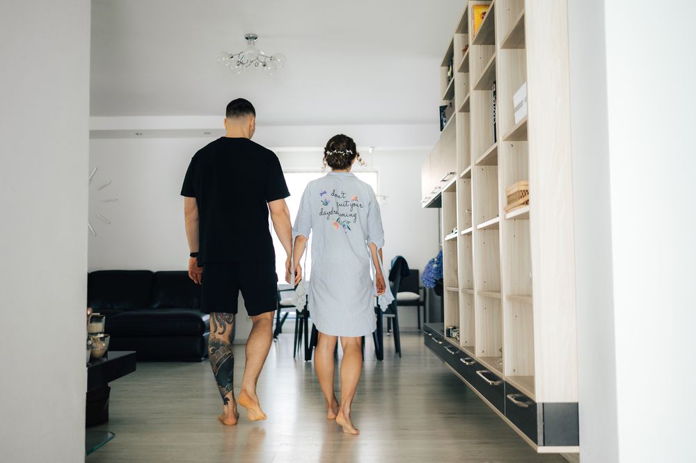 Couple holding hands walking barefoot through a modern bright living room with white walls and wooden bookshelves.