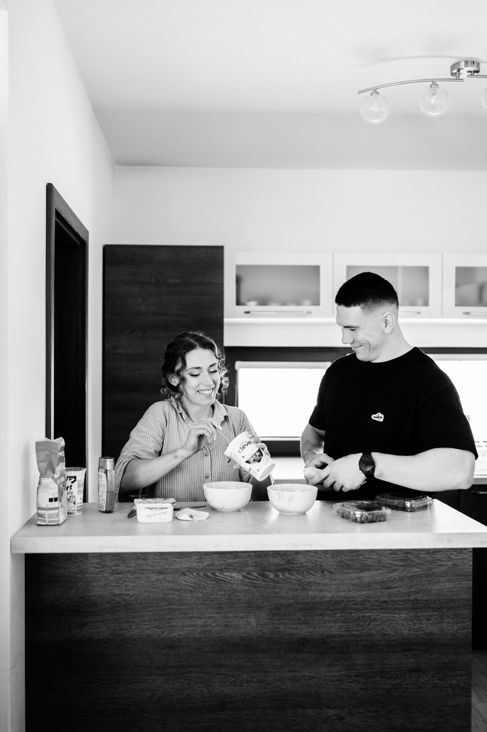 Couple smiling and preparing food together at a modern kitchen counter in black and white photo.