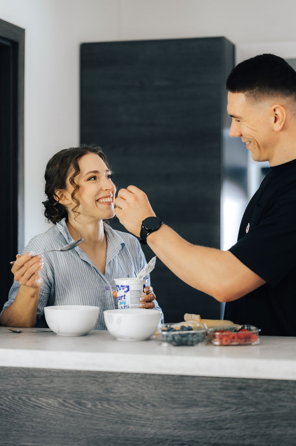 A smiling couple sharing breakfast at a kitchen counter, man feeding woman yogurt in a bright modern home.