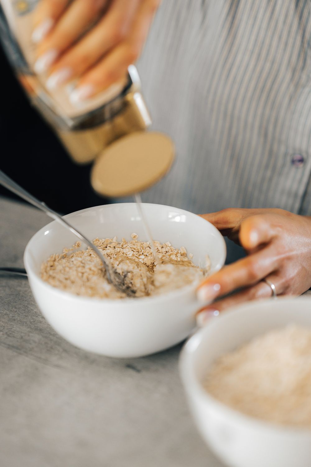 Person pouring milk into a white bowl of oatmeal with granola toppings on a gray countertop.