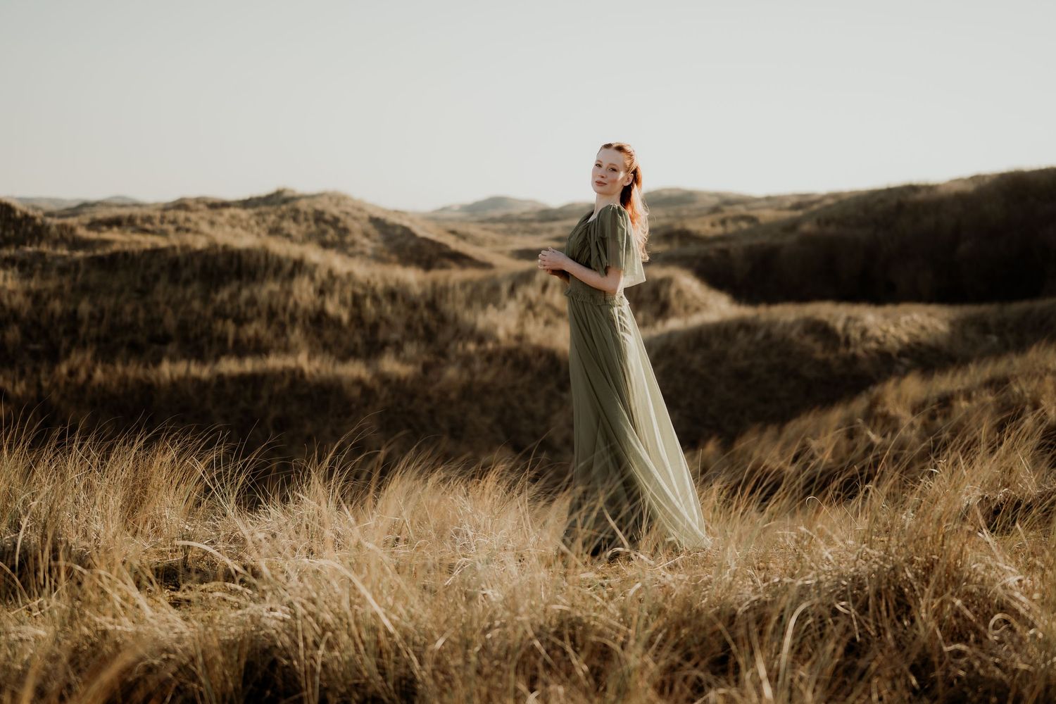 Woman in flowing sage green dress standing among golden grass dunes, gazing upward in a serene outdoor landscape.