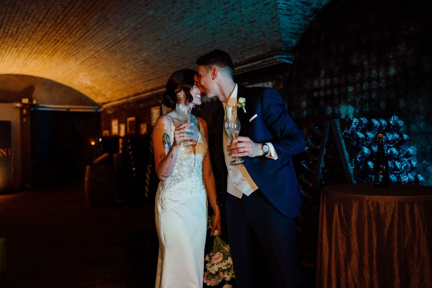 Bride and groom sharing a romantic kiss while holding champagne glasses in dimly lit venue.