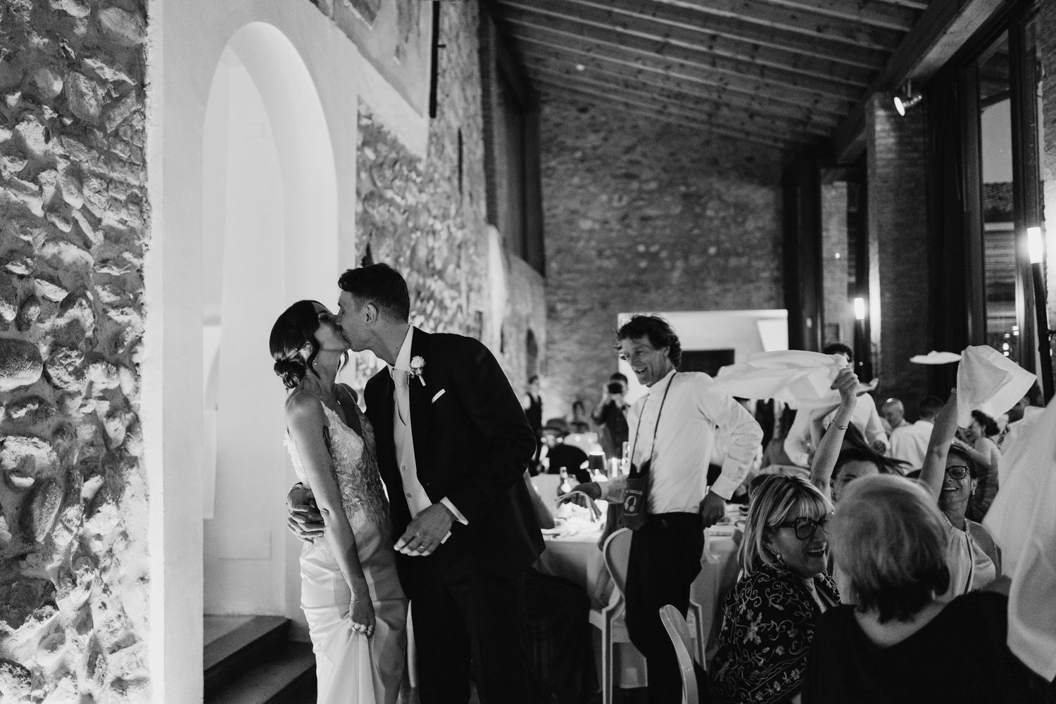 Bride and groom share a kiss during their wedding reception in an elegant stone venue with guests celebrating around them.