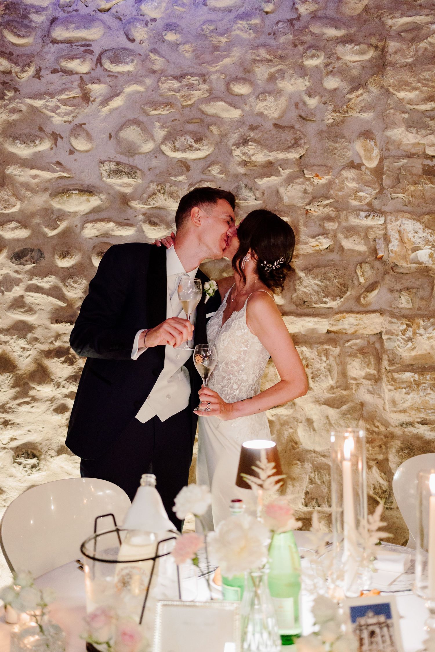 Newlyweds share a romantic kiss holding champagne glasses against a rustic stone wall at their candlelit wedding reception.