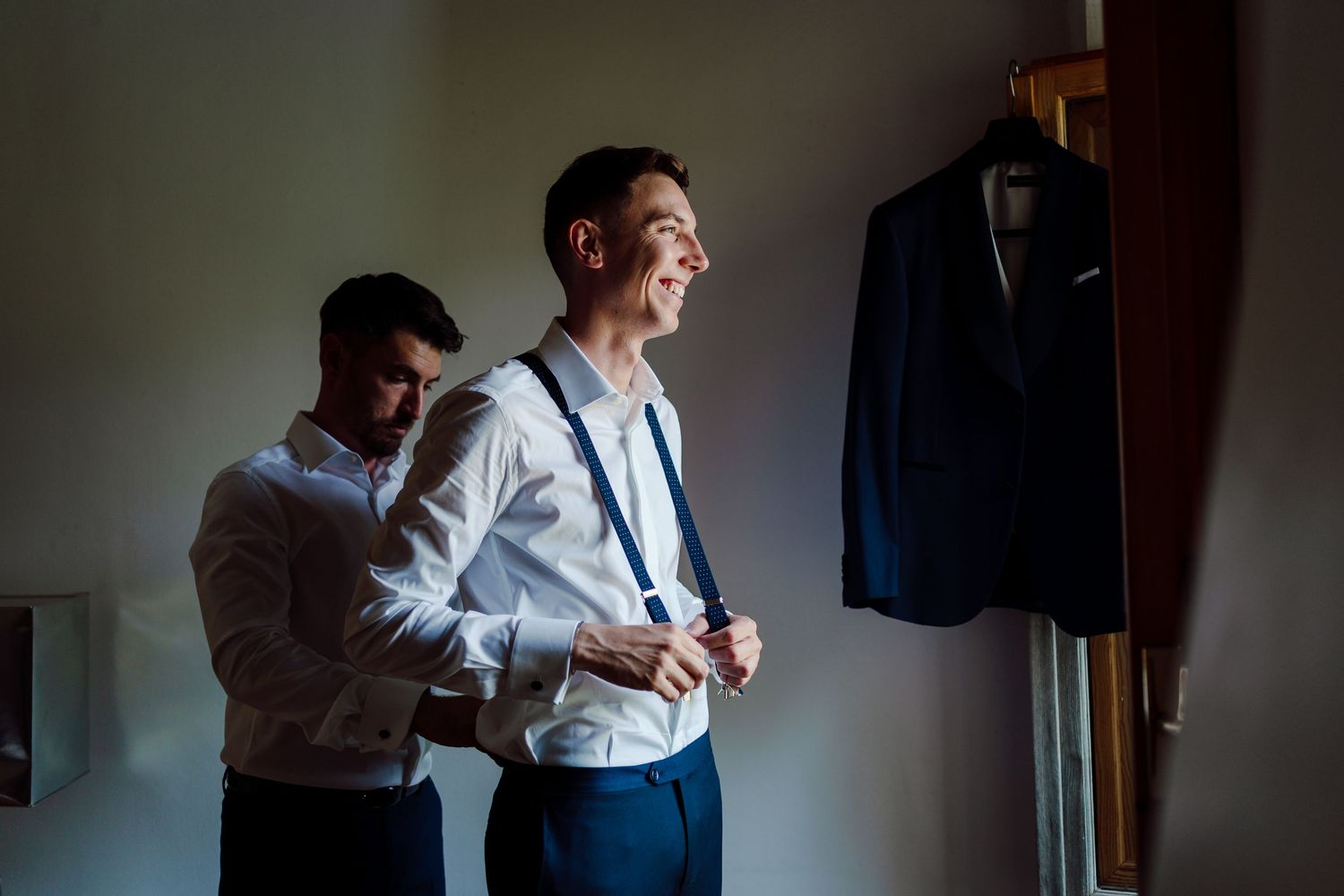 Groomsman helping smiling groom with suspenders while navy wedding suit hangs on wooden wardrobe.