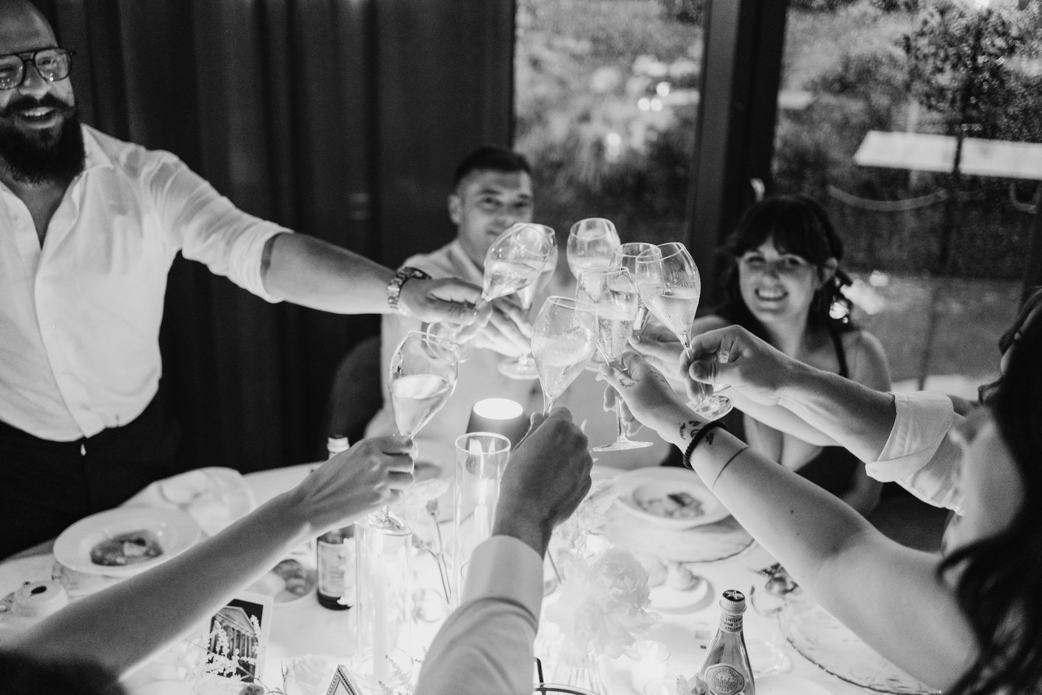 Wedding guests raise their glasses in a celebratory toast at a festive reception dinner table with bottles and glassware.