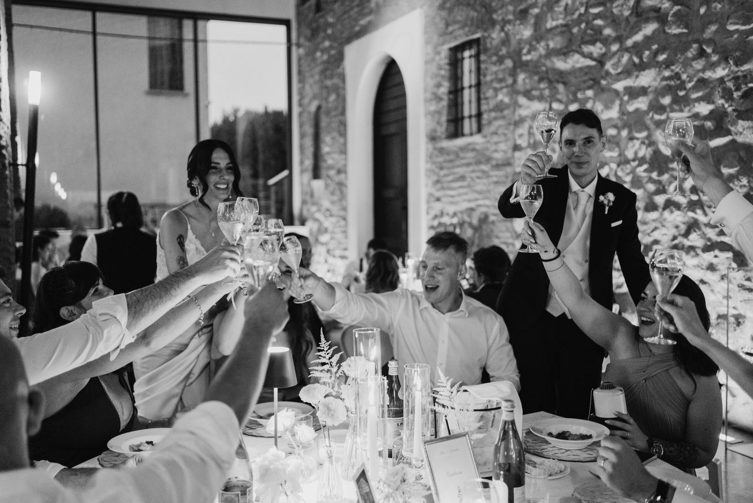 Bride, groom, and wedding guests enthusiastically raise glasses in a joyful champagne toast at the reception dinner.