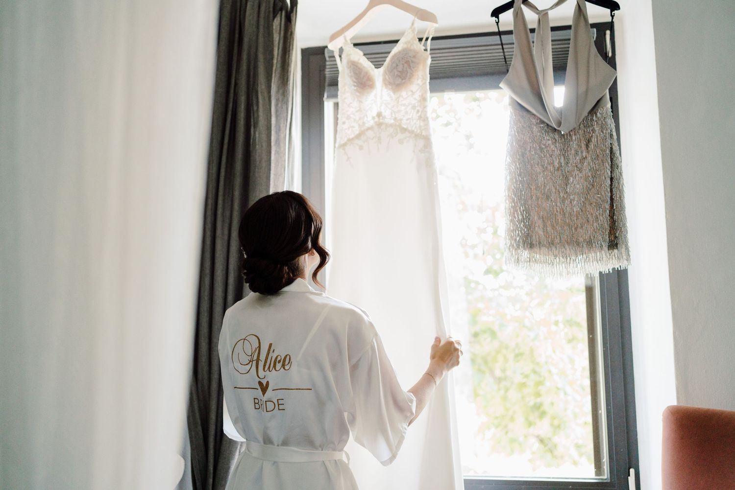 Bride in personalized white robe views her wedding dress hanging by a bright window before ceremony.