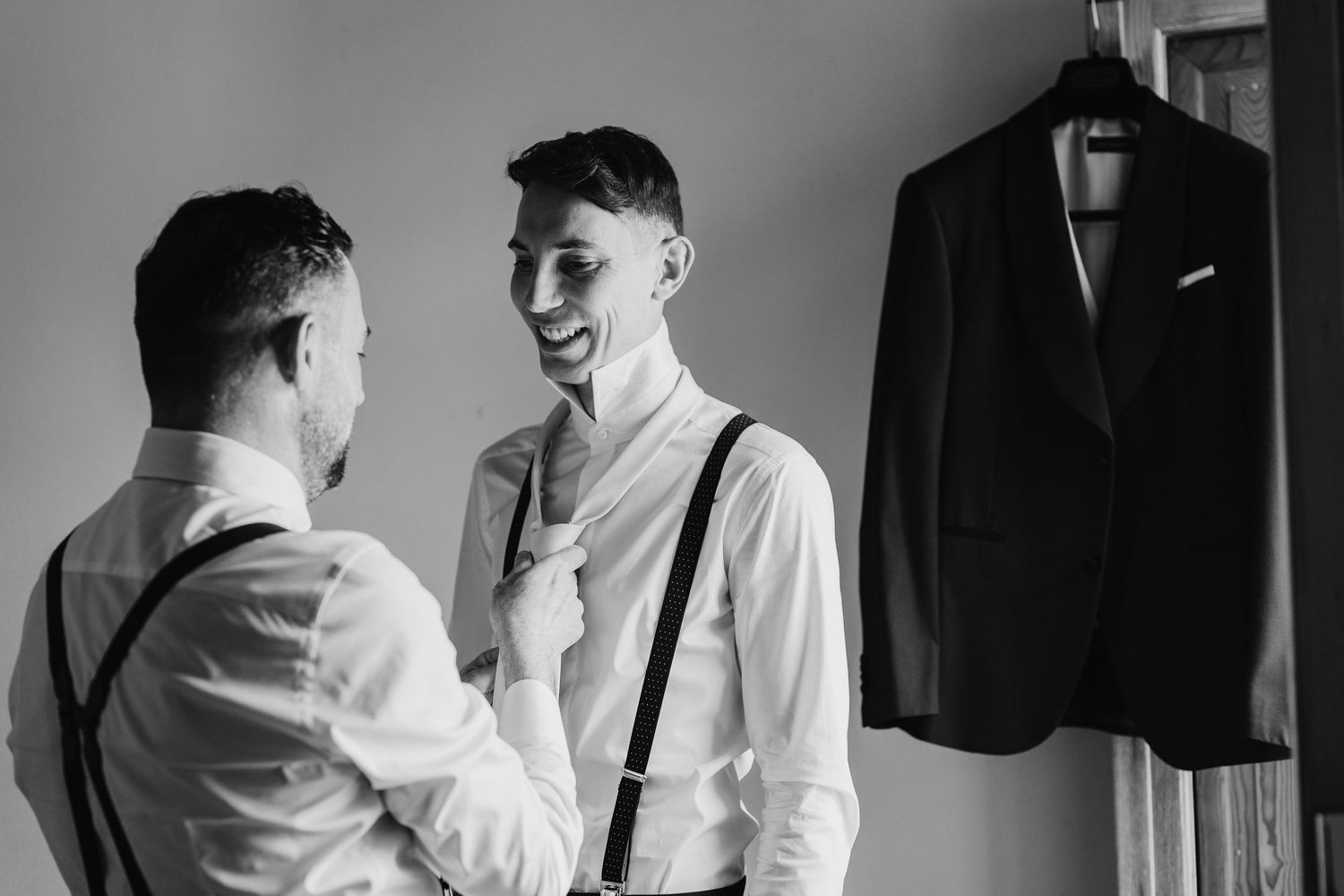 Black and white photo of two groomsmen in suspenders laughing while adjusting tie before wedding.