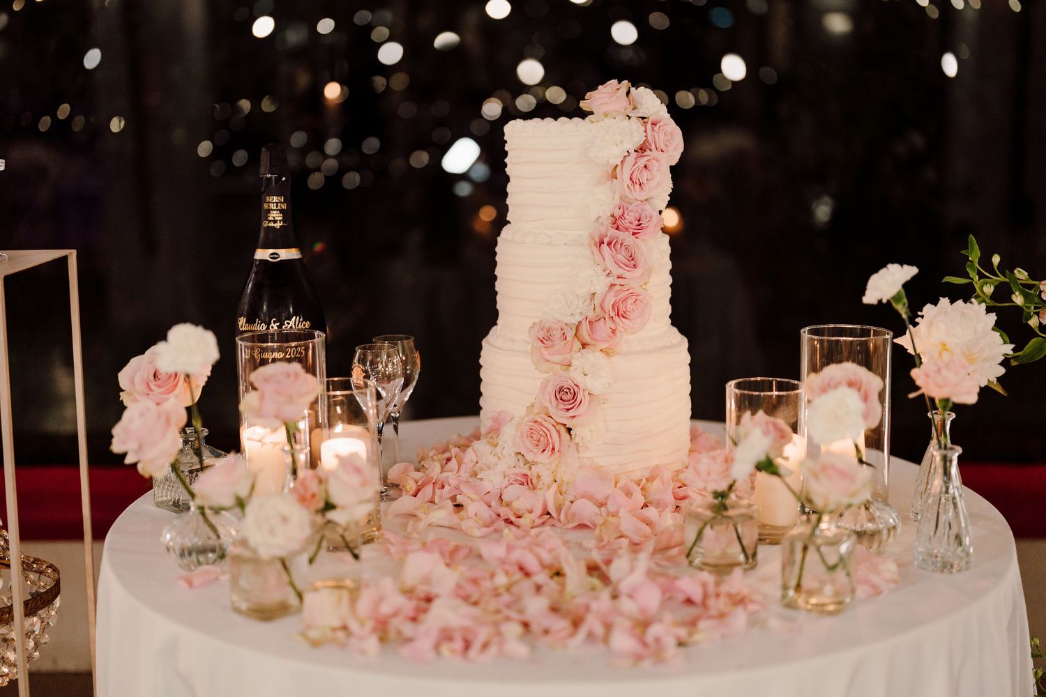 Elegant three-tier white wedding cake adorned with pink roses surrounded by candles and floral arrangements on a draped table.