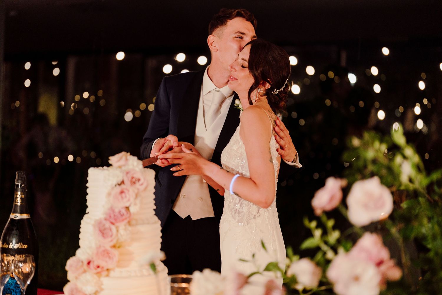 Bride and groom share a tender kiss while cutting their beautiful white and pink floral wedding cake at reception.