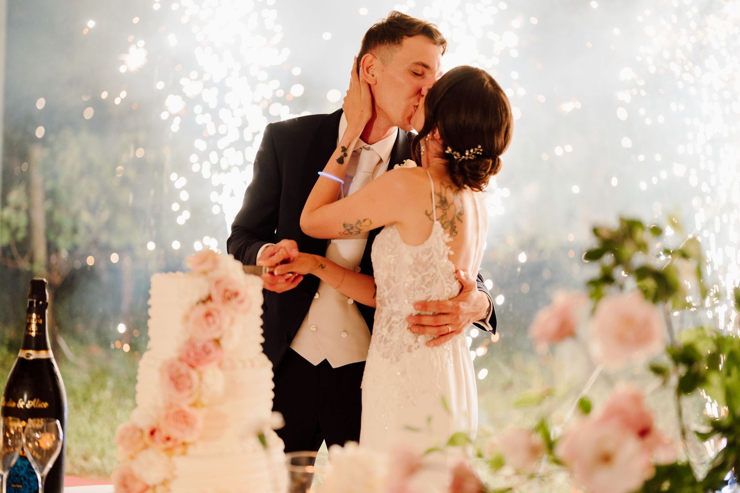 Newlyweds kiss romantically beside their elegant pink floral wedding cake with sparkling lights glowing in the background.