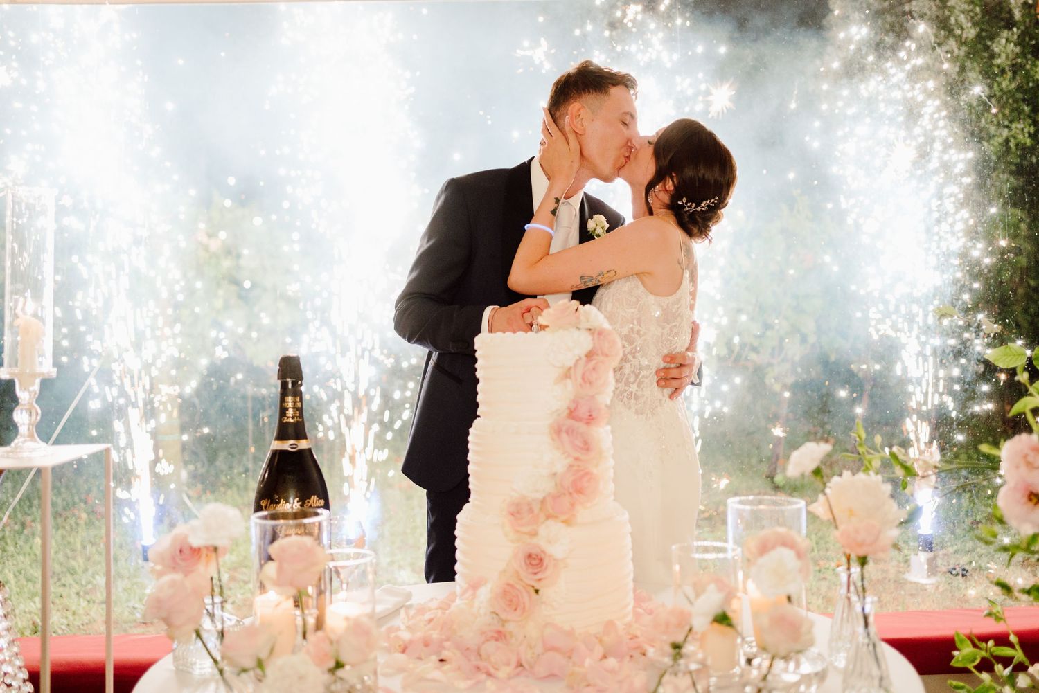 Bride and groom share a passionate kiss in front of their stunning wedding cake with dazzling sparkler fountains behind them.