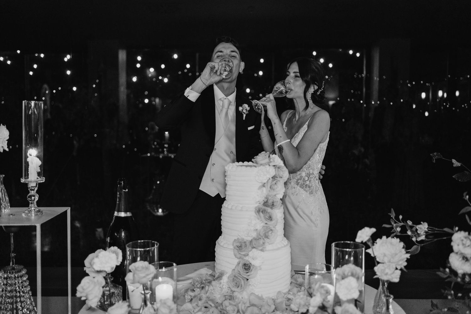 Bride and groom laughing while cutting their elegant tiered wedding cake at a romantic evening reception.
