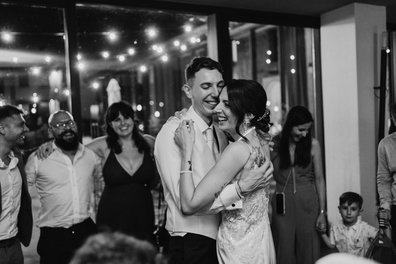 Bride and groom laugh together during their first dance surrounded by smiling wedding guests.