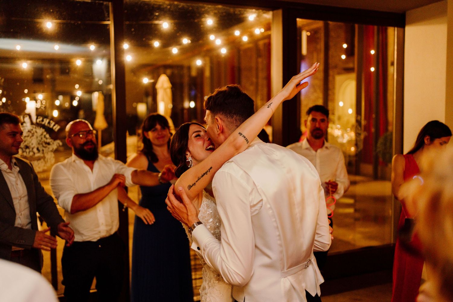 Bride reaches up joyfully during first dance with groom while guests watch at warmly lit reception.
