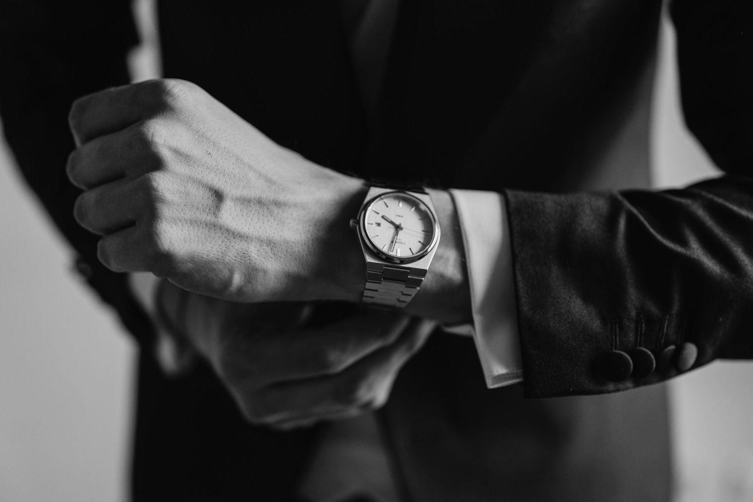 Close-up black and white photo of groom adjusting elegant watch cufflink on wedding suit sleeve.