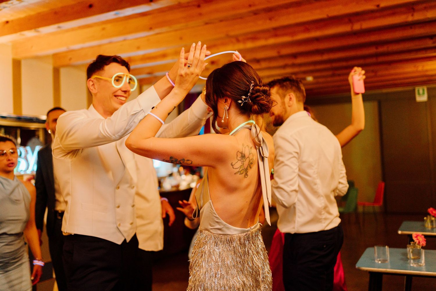 Couple wearing novelty glasses dance energetically with tattooed guest at a festive wedding reception.