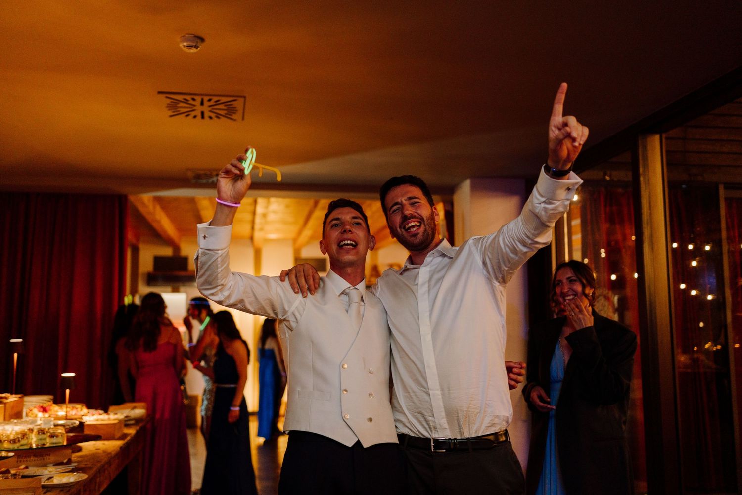 Two joyful groomsmen celebrate with arms raised at a lively and fun wedding reception party.