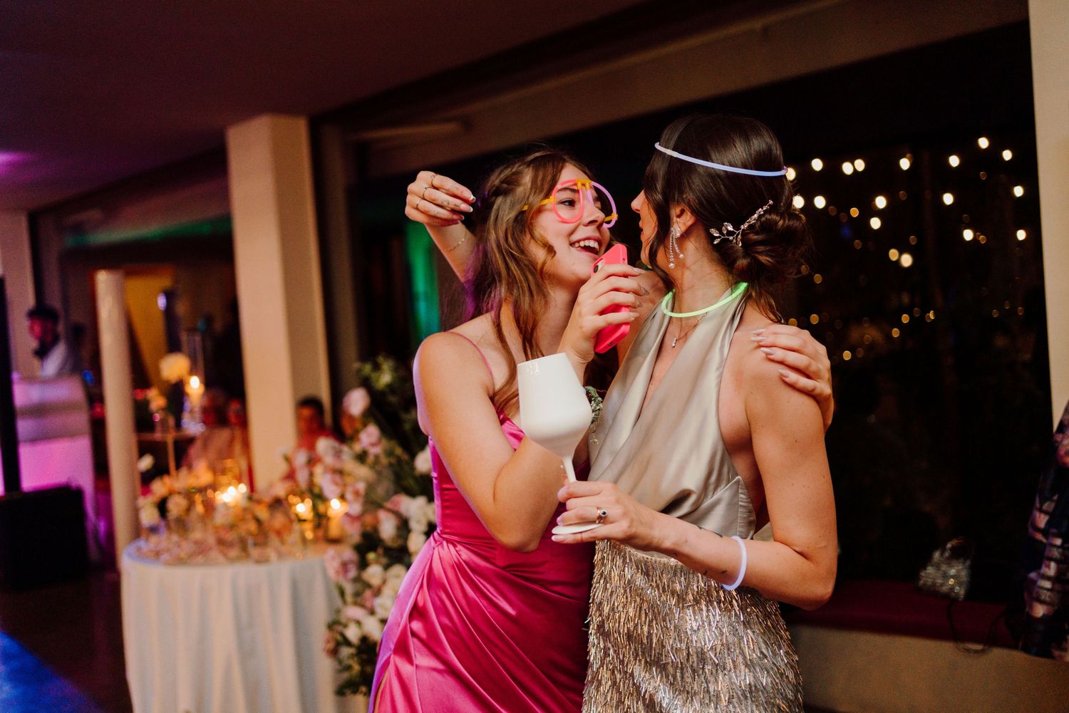 Two women laughing and dancing together at a wedding reception, one in a pink dress holding a white cocktail.