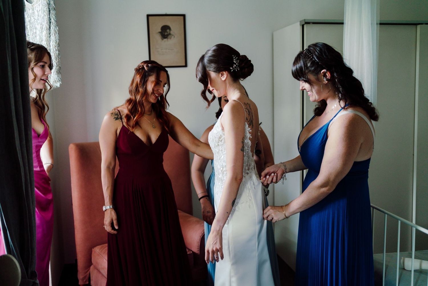Bridesmaids in colorful gowns help bride with her white lace wedding dress during bridal preparations.