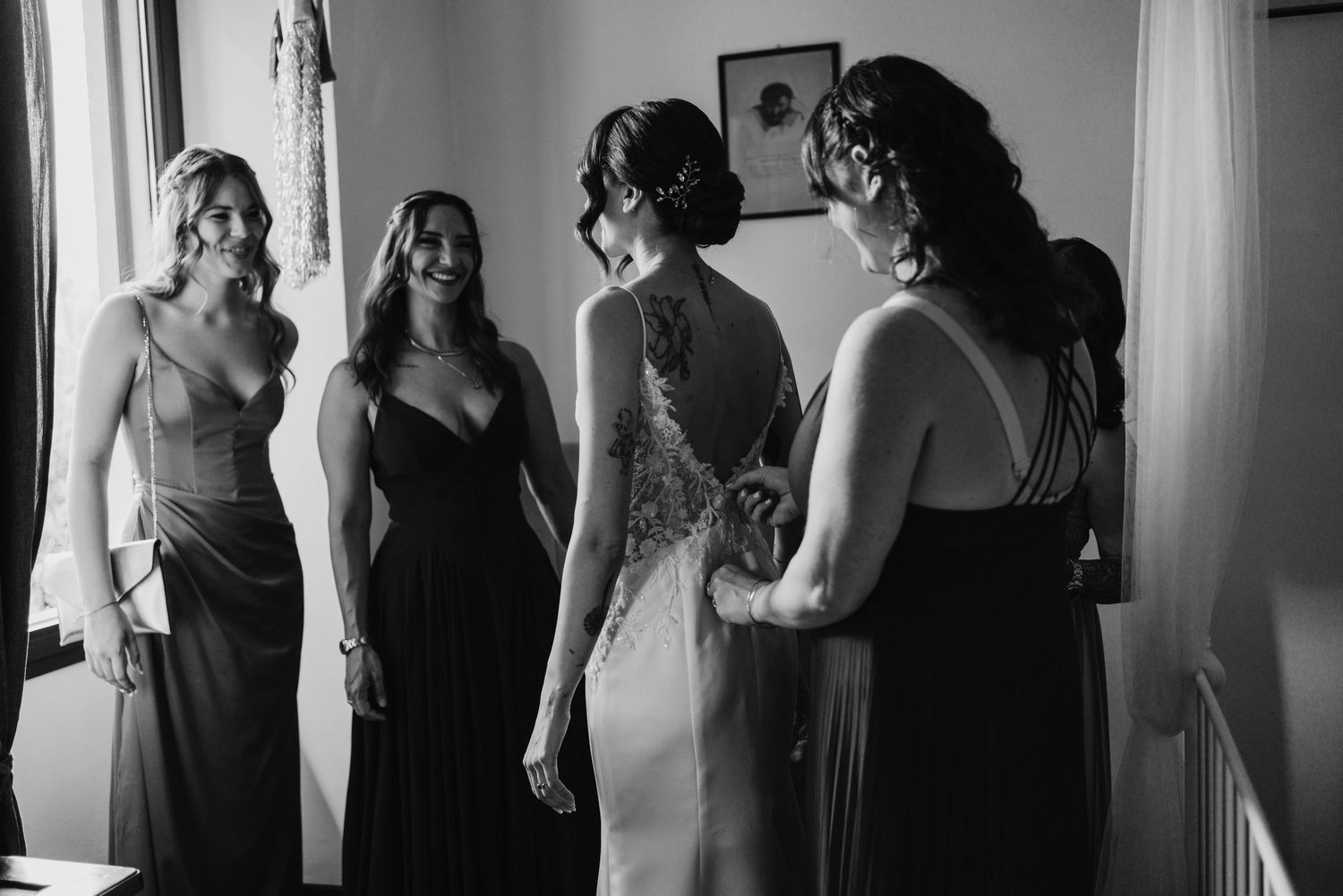 Black and white photo of smiling bridesmaids helping bride with her elegant lace wedding gown.