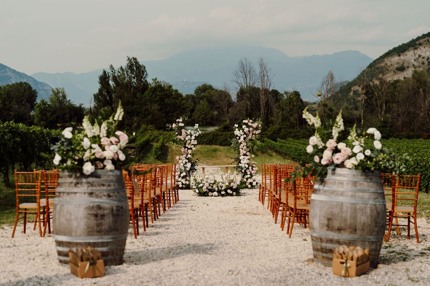 Elegant outdoor wedding ceremony aisle with wine barrels, floral arrangements, and mountain backdrop.
