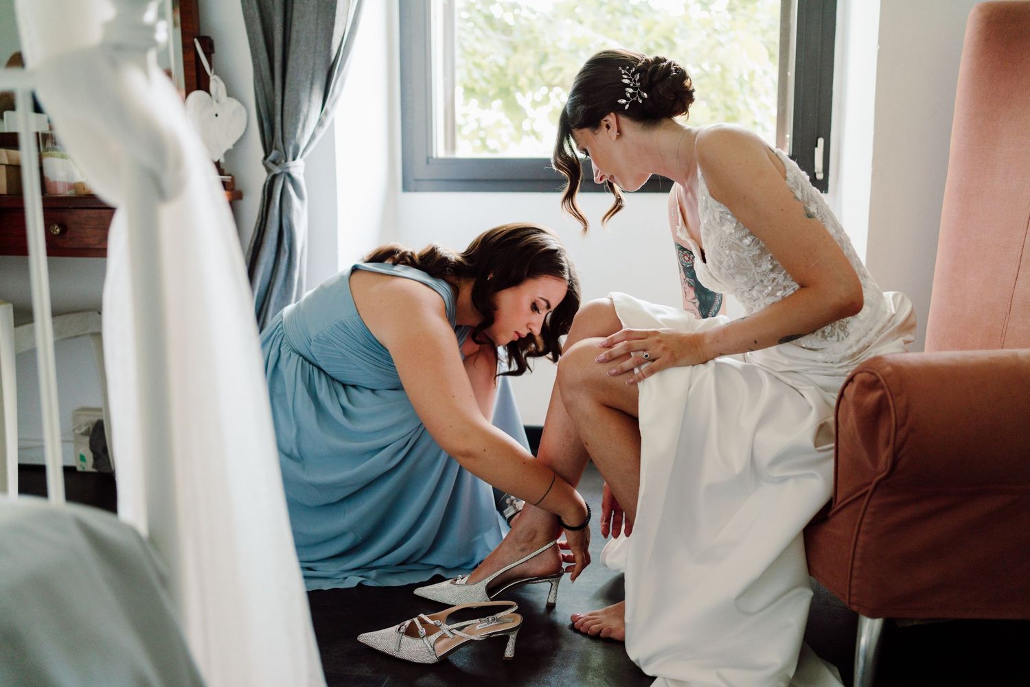 Bridesmaid in blue dress helps bride put on her wedding shoes during bridal preparations.