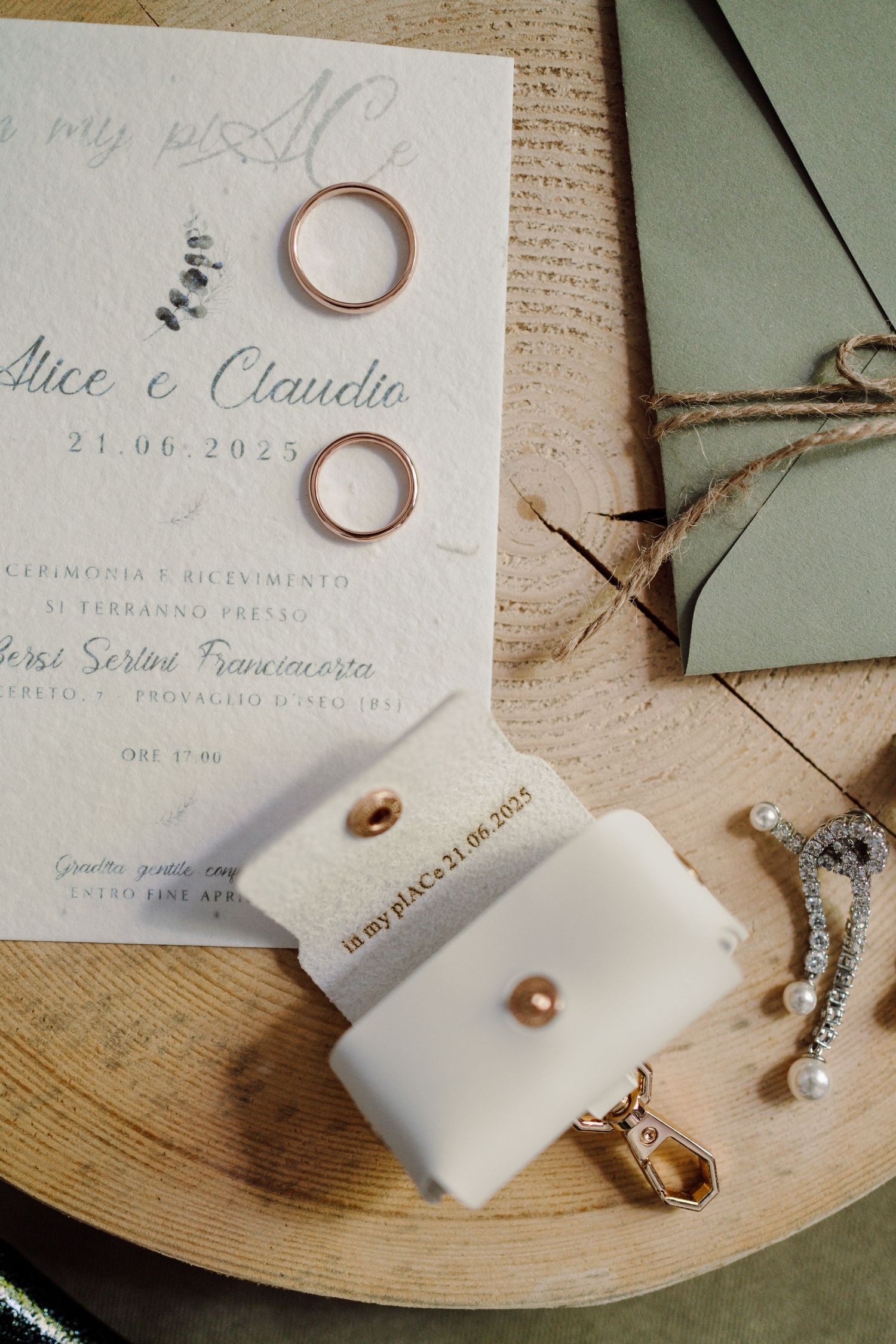 Wedding details flat lay featuring gold bands, invitation for Alice e Claudio dated 21.06.2025, pearl earrings, and sage envelope.