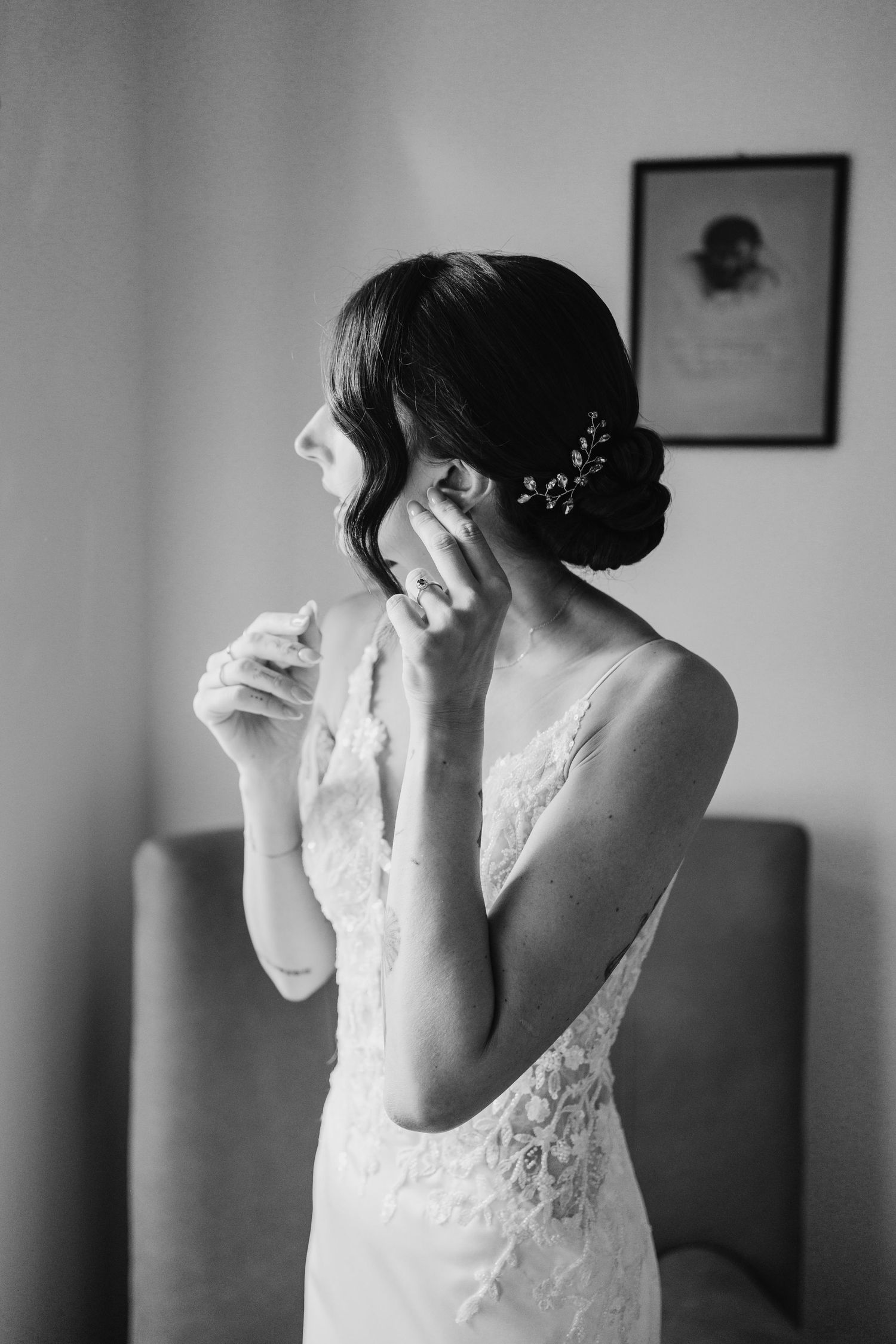 Black and white portrait of bride in lace gown putting on earrings with floral hair accessory.