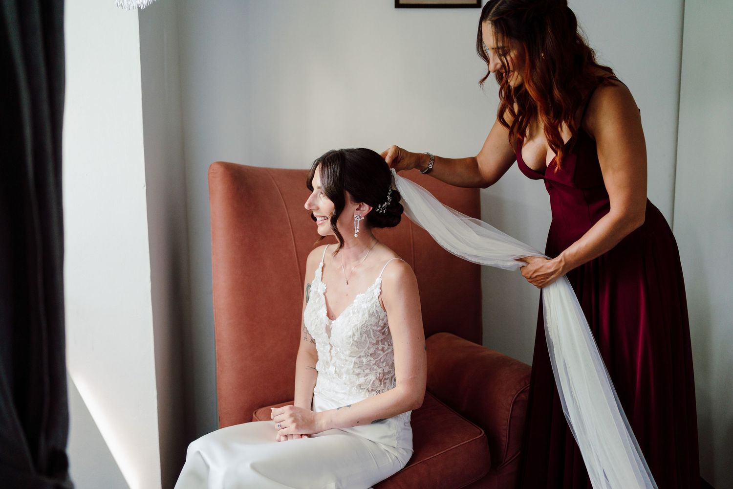 Bridesmaid in burgundy gown adjusts brides veil while she sits in a chair before the wedding.