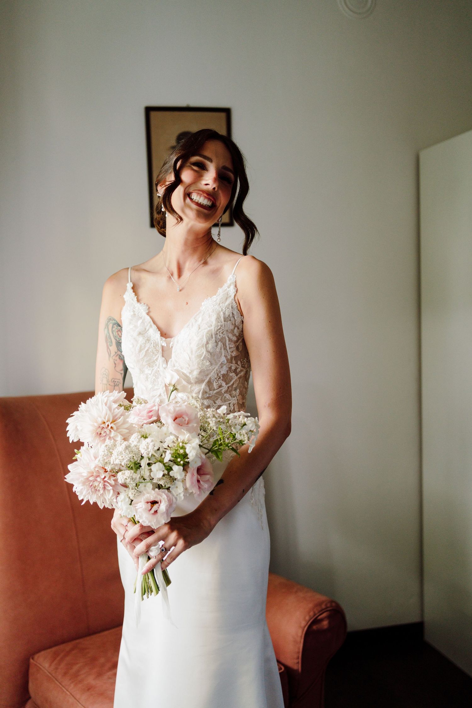 Radiant bride in lace spaghetti strap wedding gown smiling while holding a soft pink floral bouquet.