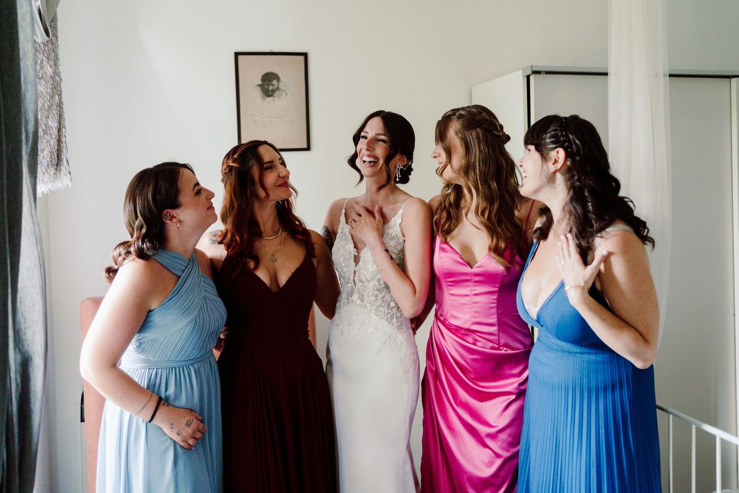 Joyful bride surrounded by four bridesmaids in colorful dresses laughing together before the wedding.