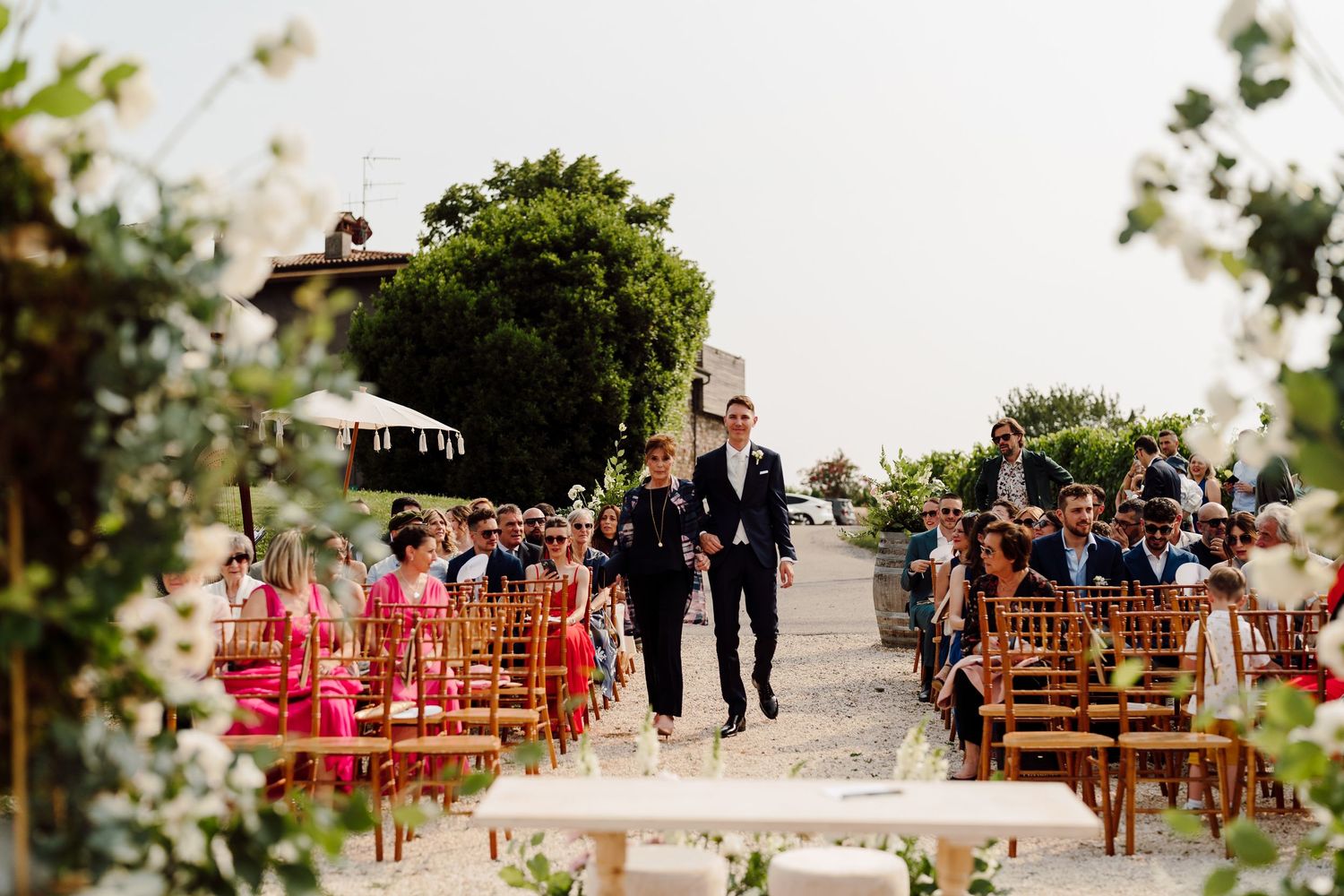 Groom walking down outdoor wedding ceremony aisle with groomsman past seated guests in vineyard.