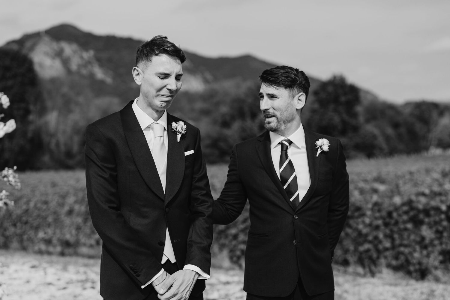 Two grooms in dark suits with boutonnieres share a tender moment outdoors at their wedding ceremony.