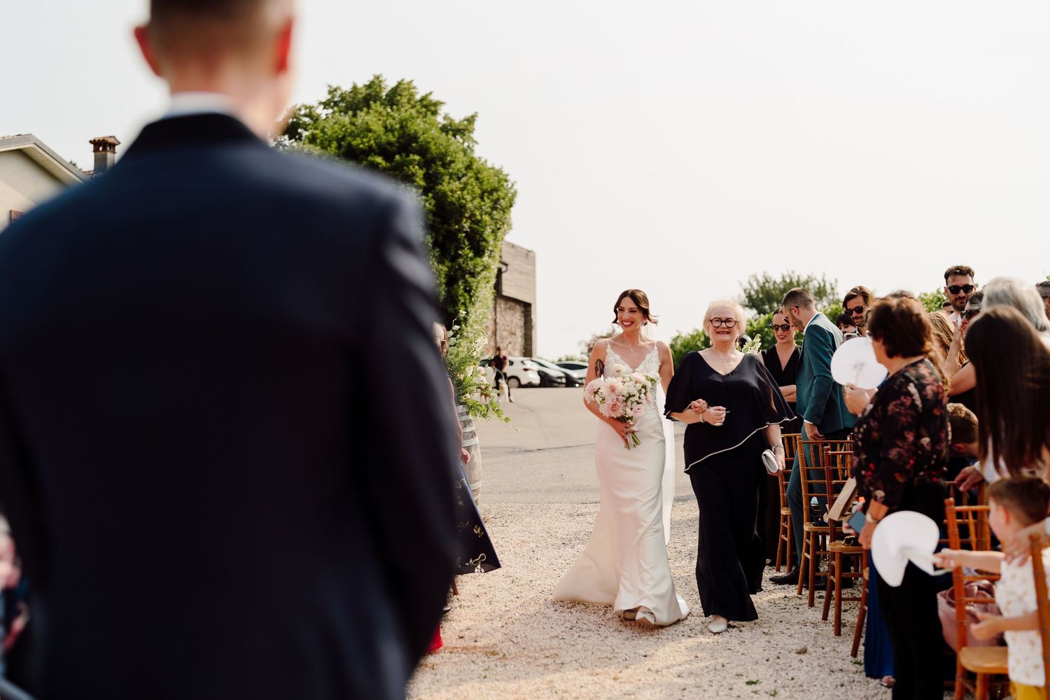 Bride walks down outdoor aisle with escort toward waiting groom at scenic vineyard wedding ceremony.