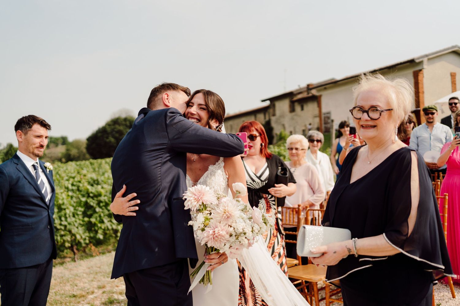 Bride in white gown embraces guest while holding pink bouquet at outdoor vineyard wedding ceremony.