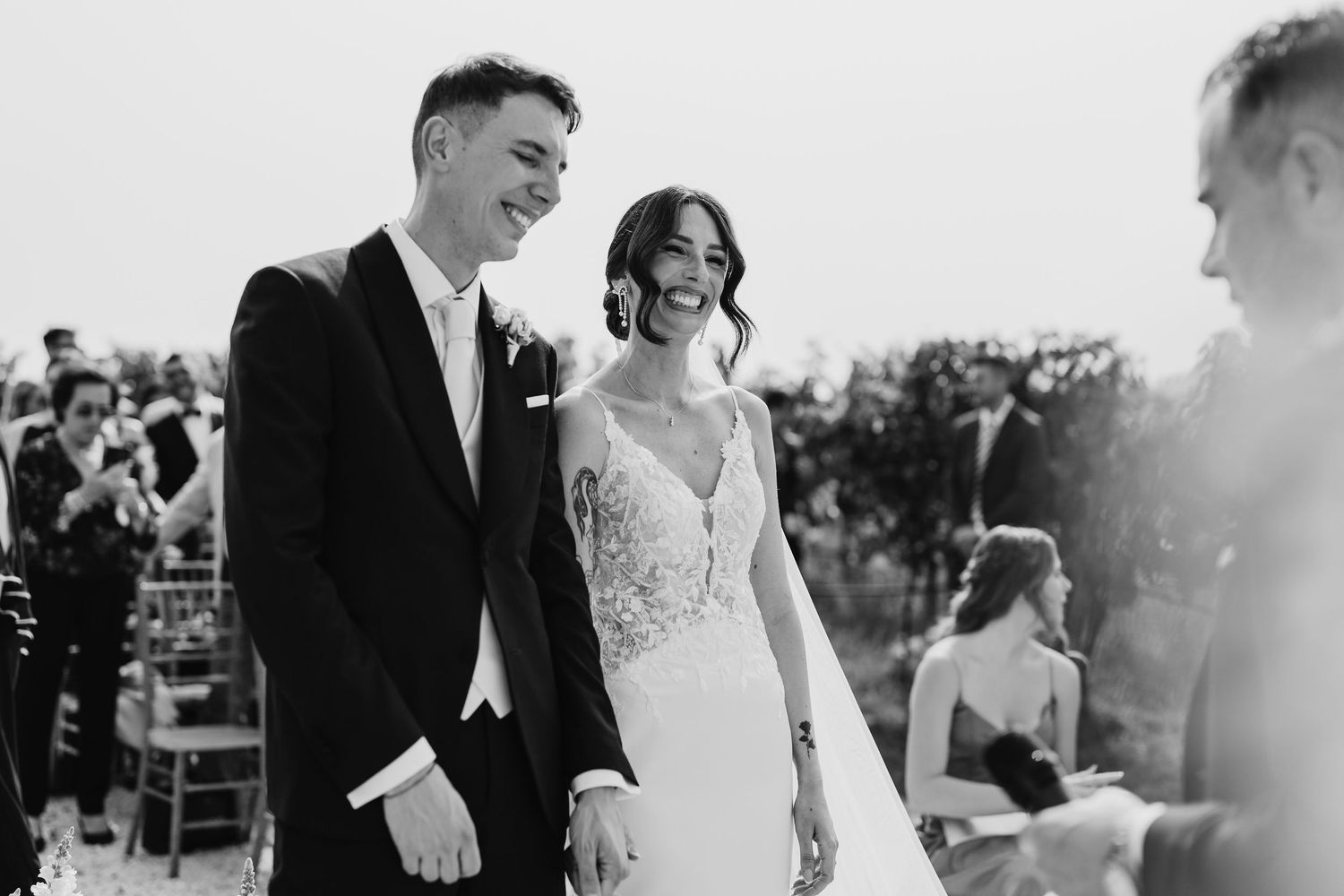 Smiling bride and groom hold hands during joyful outdoor wedding ceremony with guests in background.
