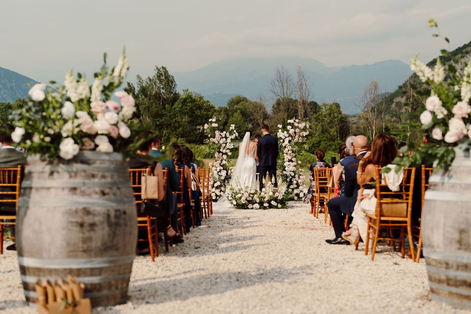 Elegant outdoor wedding ceremony aisle lined with wine barrels and white floral arrangements in mountains.