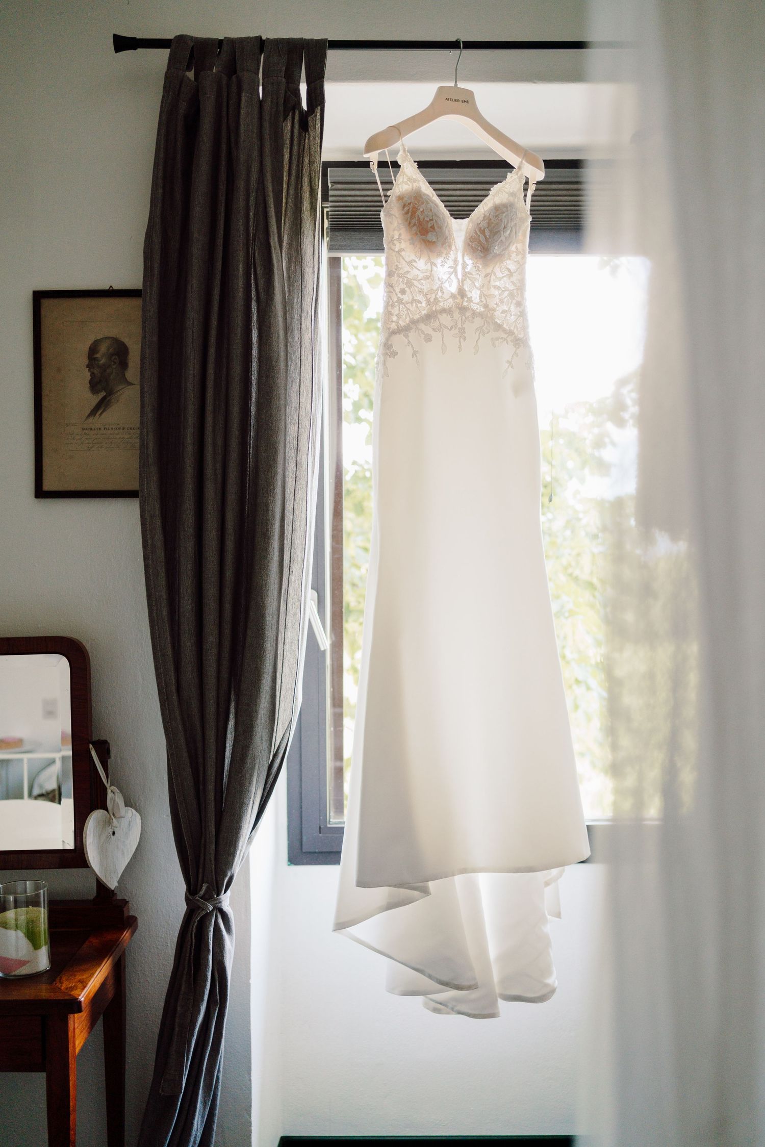 Elegant lace wedding dress hanging on wooden hanger in sunlit window on wedding morning.