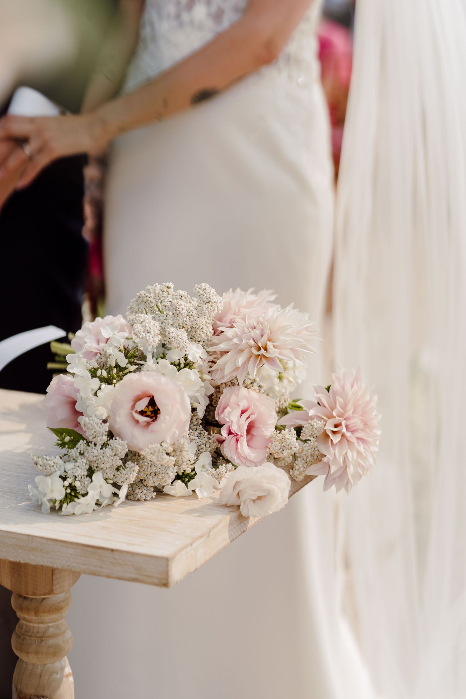 Delicate bridal bouquet of pink dahlias and white flowers rests on wooden table at wedding.