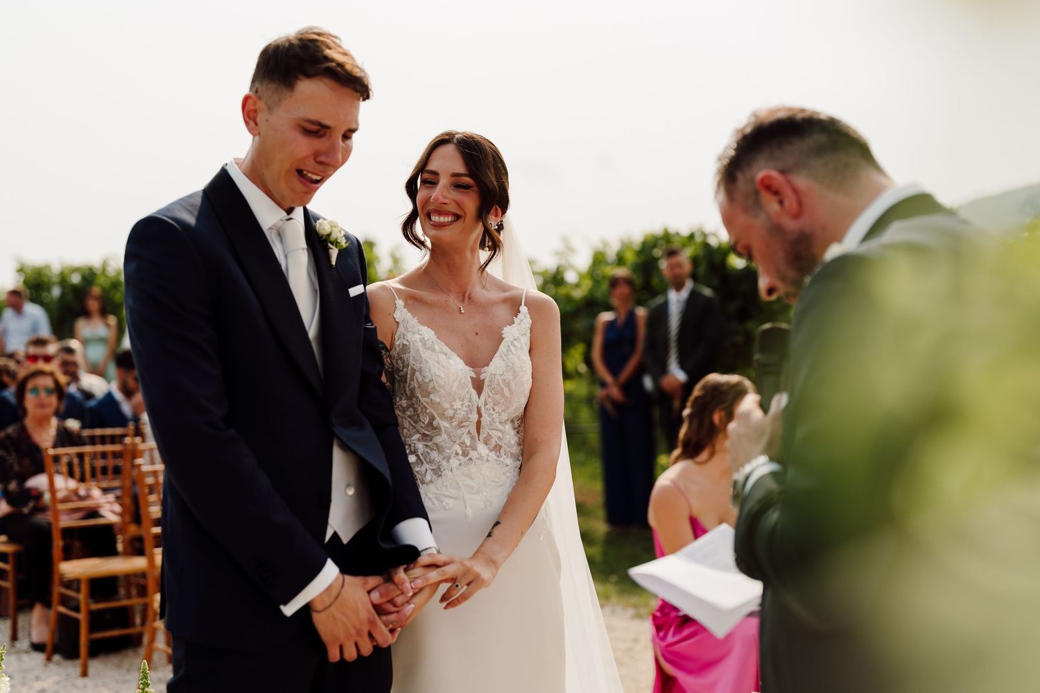 Laughing bride and groom hold hands during romantic outdoor wedding ceremony with guests seated behind.