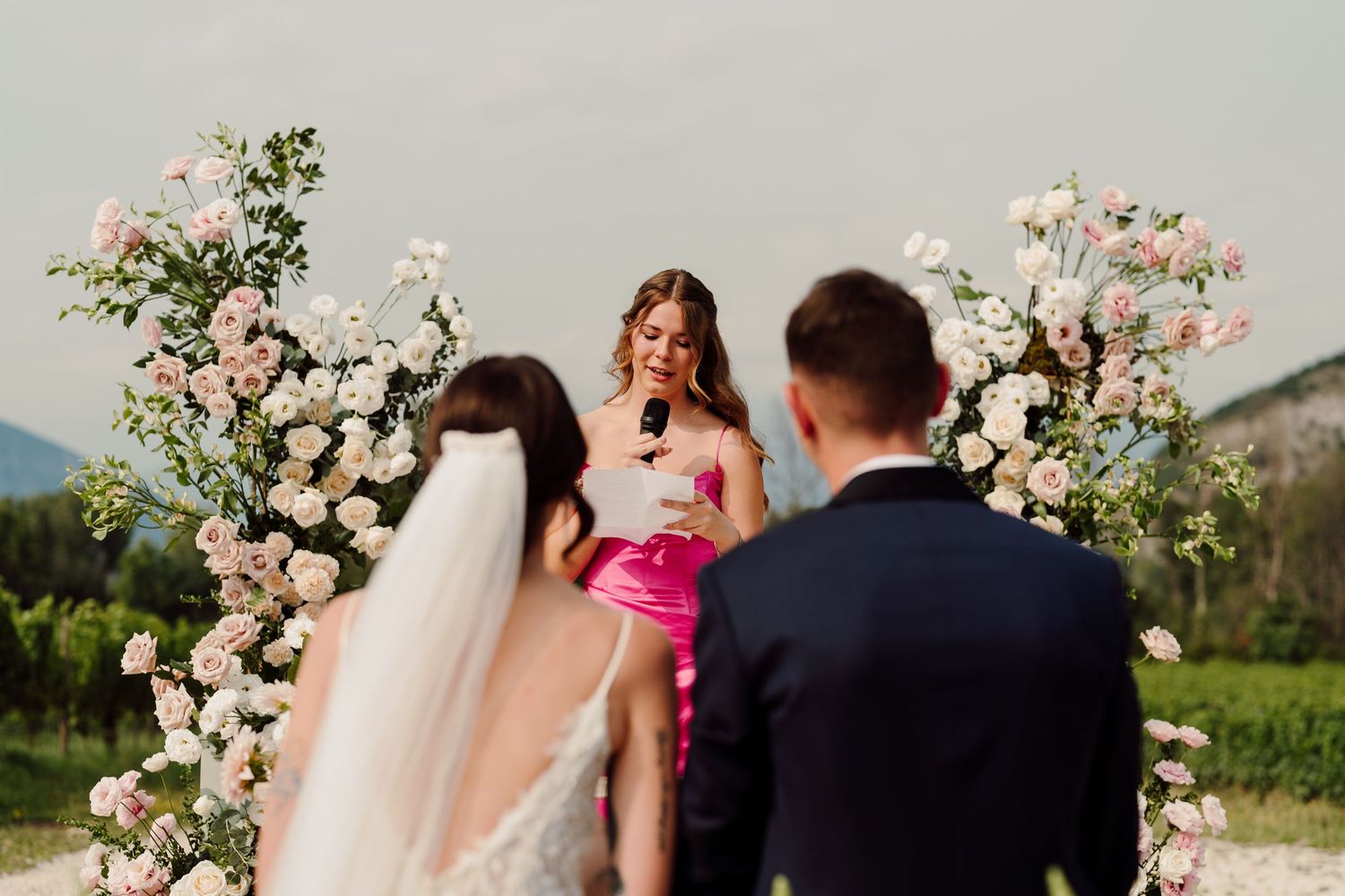 Officiant in pink dress speaks at floral arch during outdoor vineyard wedding ceremony at dusk.