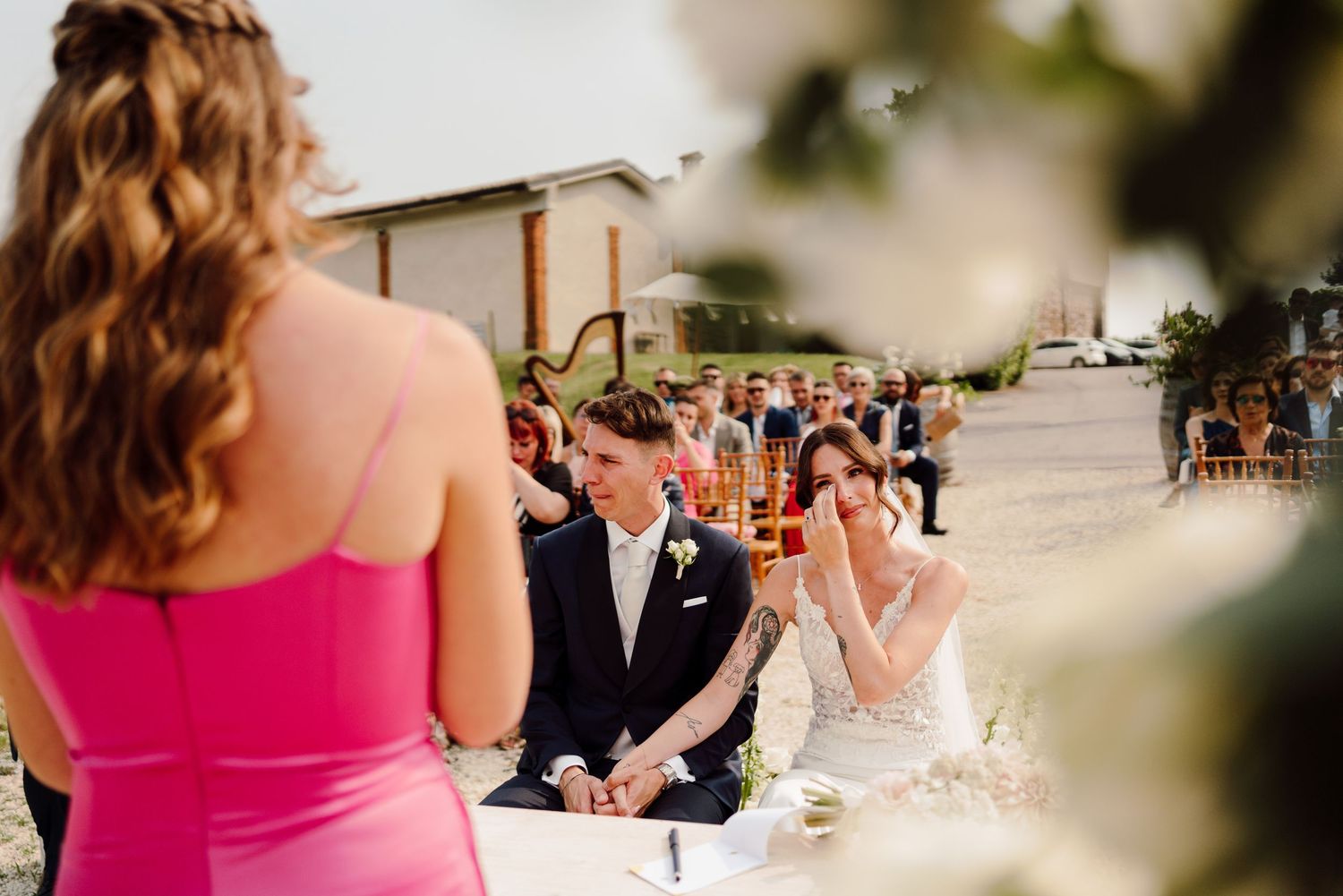 Emotional bride wipes tears as bridesmaid in pink delivers heartfelt speech at outdoor wedding reception.