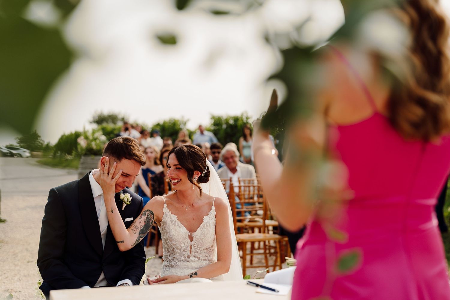 Bride and groom laughing joyfully during outdoor wedding ceremony with guests seated in background.