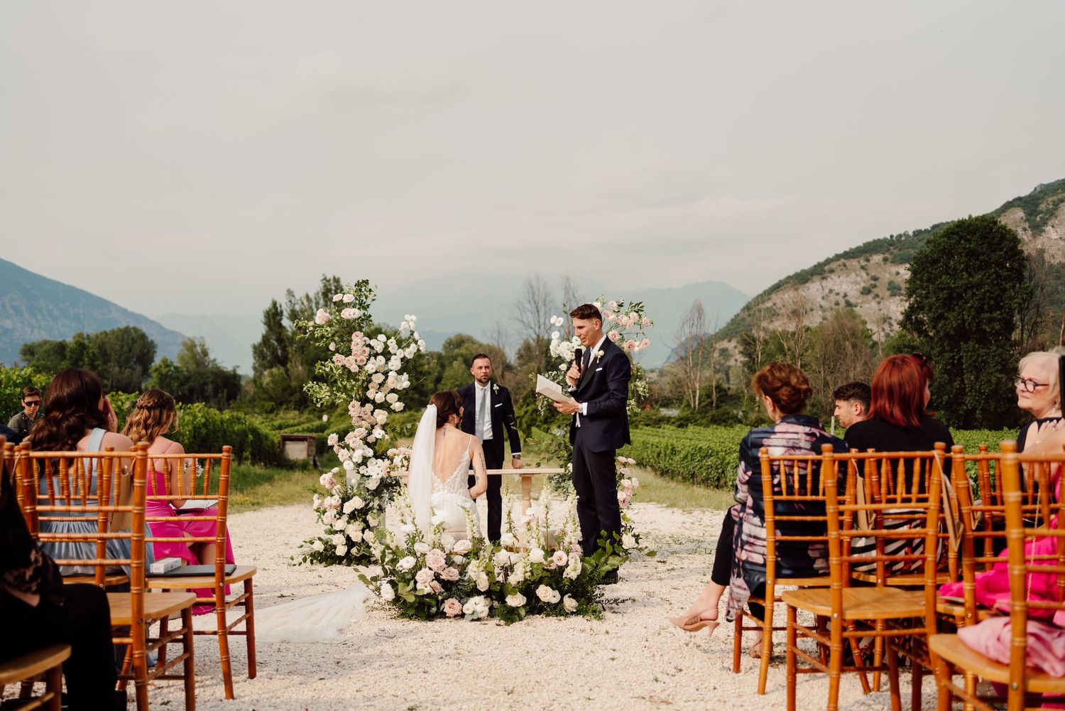 Outdoor wedding ceremony with floral arch, mountains in background, and guests seated on wooden chairs.